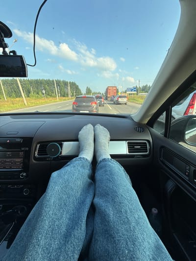 This media file depicts a first-person perspective from inside a car traveling on a road in Novoe Mart'yanovo, Russia. The scene is oriented vertically. The primary subjects are a person's legs, clad in blue denim jeans and white socks, resting on the dashboard. Their feet are visible, contributing to a casual and relaxed atmosphere.

Outside the car, the view through the windshield shows a bright, sunny day with a clear blue sky and scattered white clouds. Traffic is visible on the road ahead, suggesting the car is part of a convoy or experiencing a traffic jam. A variety of vehicles, including cars and a truck, are present. Roadside greenery, characterized by trees and tall grass, lines the highway, with what appear to be utility poles or streetlights interspersed. A blue road sign with Cyrillic text is also visible, indicating a location in Russia.

Notable details include the presence of a dashcam mounted on the windshield, positioned in the lower left of the frame. The car's interior is partially visible, including the dashboard with air vents and a car stereo system displaying the time "19:14" and radio station information, suggesting it's evening. The overall mood is one of a typical road trip, with the person in the car opting for a comfortable posture. The weather appears pleasant, conducive to travel.