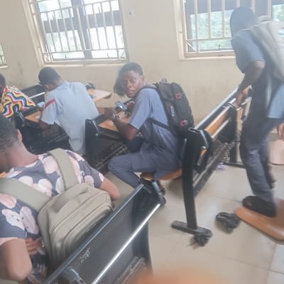 The image shows a group of young people, likely students, in a classroom or lecture hall. The scene is captured from a slightly elevated perspective, looking down at the subjects. Most of the individuals appear to be wearing casual clothing, with some carrying backpacks. One person is prominently featured in the foreground, wearing a light blue shirt and dark trousers, with a black backpack slung over their shoulder. They are looking towards the camera with a neutral expression.
The setting appears to be an indoor space with windows that allow natural light to enter, suggesting it could be daytime. The windows have a grid-like structure. Desks and chairs, typical of a learning environment, are visible, and some are partially occupied or empty. The floor is tiled. There are also some black plastic bags scattered on the floor in the background. The image's orientation is skewed, making it appear as if the subjects are lying down or the camera is tilted.
The overall impression is one of a casual, everyday moment within an educational institution. There is no indication of a specific event or dramatic activity taking place. The lighting suggests it is daytime. There is no visible text in the image. The location context provided is Wukari, Nigeria, which aligns with the appearance of the people and the setting.