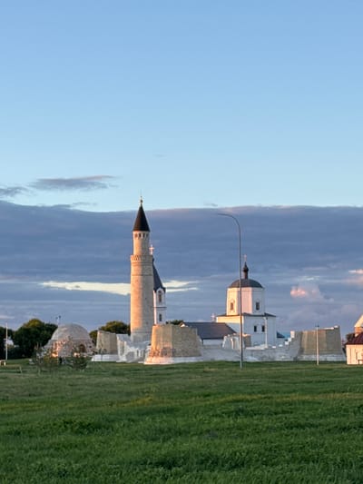 The image shows the ancient ruins of Bolgar, Russia, at sunset. In the foreground, a vast expanse of green grass stretches towards the horizon. In the middle ground, a tall, light-colored minaret with a black conical roof stands prominently. To its right, a white building with a dark dome and cross, likely a church, is partially visible. The ruins feature low, stone walls and a domed structure on the left. The sky is a gradient of light blue at the top, transitioning to darker, dramatic clouds tinged with pink and purple towards the horizon. A single lamppost stands in front of the white building. The setting sun casts a warm glow on the minaret and the ruins. There are no people clearly visible in the image.