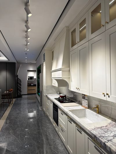 This is an interior shot of a modern kitchen, likely located in a private residence within the Chancheng District, China. The perspective is from the floor, angled upwards towards the ceiling and kitchen cabinets.
The kitchen features sleek, white cabinetry with gold hardware. Some upper cabinets have recessed lighting. Countertops are marble, possibly white marble with gray veining, extending along the wall with a gold-toned faucet and integrated sink. A stovetop is visible on the counter, with a range hood above.
Below the cabinets are drawers with horizontal gold handles. Appliances such as an oven and possibly a dishwasher are built-in. The flooring is grey marble-like tile with prominent veining.
The scene extends beyond the kitchen into other rooms. A hallway leads towards a garden view with potted plants, suggesting an outdoor area. In the foreground is a staircase with wrought iron railings and some modern art pieces. The walls are painted in a neutral white or off-white tone.
Along one wall, there is track lighting with several cylindrical spotlight fixtures. The lighting illuminates the space and highlights the modern design aesthetic. No people are visible.