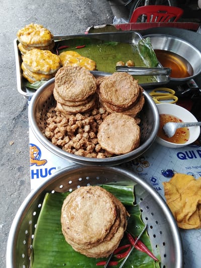 A street food stall in Phan Thiết, Vietnam displays an assortment of fried fish cakes. In the foreground, a stainless steel colander holds a stack of round, golden-brown fried fish cakes resting on a bed of banana leaves. Beside this colander, another stainless steel bowl is filled with smaller, nugget-shaped fried fish pieces. To the right, a small white bowl contains a dipping sauce with visible chili pieces, and a metal spoon is partially submerged in it. Behind these, a shallow metal pan holds a small amount of cooking oil. Further back, a tray contains several larger, flatter fish cakes, some appearing slightly greased, also on banana leaves, with two red chilies on the side. Yellow kitchen tongs rest on the tray. In the background, a red plastic chair is visible. The scene is set outdoors on a pavement, with the ground texture suggesting a street.