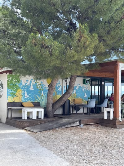 The scene shows a cafe located at coordinates 43.445, 16.637 in Dugi Rat, Croatia. A large pine tree provides shade over a wooden patio. The patio is connected to a building with a mural depicting a nautical scene with sailboats and a woman holding a blue bag. Two white, square tables with cushioned benches are located on the patio. Two people are seated at tables in the background, with a black trash can near the wall. The patio's wooden floor has a small wooden ramp leading to the gravel ground. The cafe has a white awning and windows leading to a view of the ocean in the background. The building is painted in light blue and white.