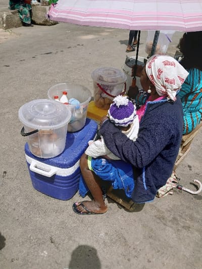 An outdoor street vending scene in Masvingo, Zimbabwe, features a woman seated on a low wooden crate, facing away, holding an infant. The woman wears a dark blue long-sleeved top, a white headscarf with red script, and black flip-flops. Her right ear has a white beaded earring. The infant wears a white garment and a purple knitted hat with a white pom-pom.

Above the subjects, a light pink and white striped umbrella provides shade. To the woman's left, a blue and white cooler rests on the ground, topped by a clear plastic bucket containing a clear liquid or ice. Adjacent to the cooler, another clear plastic bucket holds several small white bottles, possibly containing beverages, and other items. A third clear plastic container, positioned on a yellow surface, holds multiple light brown, spherical baked goods.

A second individual, wearing a blue and white patterned garment, is partially visible to the right, also seated. The ground is a grey, paved surface, resembling worn concrete or asphalt, with minor marks. A folded umbrella with a curved white handle is visible on the ground near the main woman's feet.