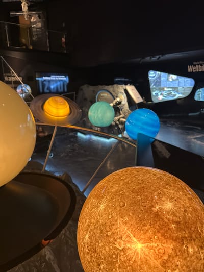 The image captures an indoor exhibit, likely a museum, in Salzburg, Austria, showcasing models of planets from the solar system. In the foreground, a large, illuminated model of a planet, possibly Venus or Mercury due to its cratered, orange-brown surface, dominates the lower right of the frame. To its left, a larger, unlit pale yellow sphere is partially visible. Behind these, several other planet models are displayed. One is a ringed planet, likely Saturn, illuminated with a warm yellow glow. Adjacent to it is a turquoise-colored planet, potentially Uranus or Neptune, and a bright blue planet, possibly Jupiter or Neptune. The exhibit features a dark, reflective surface below the planets, which appears to be water or polished material, creating reflections of the celestial bodies and the surrounding exhibit space. In the background, exhibit displays and information panels are visible, including one with text that appears to read "Weltbild". The lighting is dim, focusing attention on the illuminated planet models, creating a dramatic and educational atmosphere. The overall scene suggests an educational installation designed to inform visitors about astronomy.