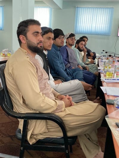 A group of men are seated at a table, likely in a meeting room, in Shurandam, Afghanistan. The men are wearing traditional Afghan clothing. The room is simple, with blinds on the windows and a table with microphones and water bottles. They appear to be engaged in a formal discussion.