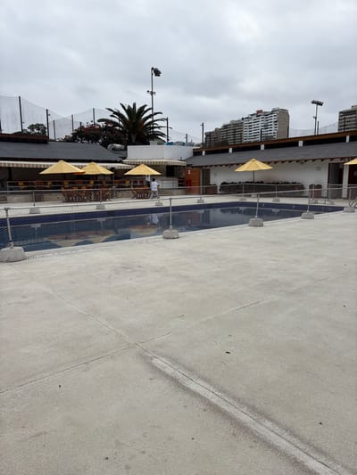 In San Isidro, Peru, an outdoor swimming pool area is depicted under an overcast sky. A dark blue rectangular pool occupies the middle ground, bordered by a light-colored concrete deck. Chain-link fencing, supported by concrete pillars, encloses the pool area. Several yellow umbrellas are open, providing shade over a seating area with tables and chairs situated behind the fence. A single person, wearing a white shirt and dark pants, walks along the paved area adjacent to the pool. In the background, a palm tree stands tall, and beyond that, several multi-story buildings are visible, characteristic of urban architecture. The overall scene suggests a recreational or club facility.