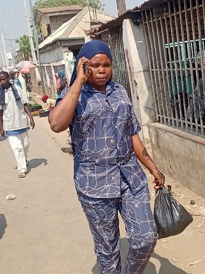 A woman in blue patterned pajamas and a headscarf stands on a dusty street, talking on a cell phone. She holds a black garbage bag. Two men walk nearby, one in a striped shirt and white pants. Behind them, a building with a metal grill overhangs part of the street. Other buildings and people are visible in the background. The scene is brightly lit.