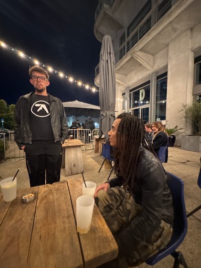 This sideways image captures an outdoor evening scene, likely on the patio or terrace of a bar or restaurant in Pantin, France. The overall lighting suggests nighttime, with string lights providing ambient illumination against a dark sky.

In the left foreground, a man with short dark hair and glasses wears a black t-shirt featuring a white circular logo that resembles a stylized "K" or an arrow, beneath a black leather jacket and dark pants. He is looking directly towards the camera.

To the right, a woman with long dreadlocks, dressed in a black leather jacket and camouflage-patterned pants, leans over a rustic wooden table. Her hands are near the table's surface, and she appears to be looking down at something.

The wooden table, which occupies a significant portion of the foreground and mid-ground, holds several opaque white plastic cups, some containing a pale yellow liquid, and all fitted with dark straws. An ashtray filled with cigarette butts is also visible on the table. Partially visible blue plastic chairs are situated around the table.

In the background, a large, grey, folded patio umbrella stands upright. Behind it, several indistinct figures of other patrons are visible in a slightly brighter, covered area. A multi-story concrete or stone building with large windows, reflecting interior lights, forms the backdrop. A potted plant can be seen near the building, adding a touch of greenery to the urban environment. The scene depicts a casual social gathering under artificial lights.