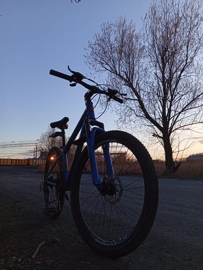 A bicycle is silhouetted against the early morning sky and landscape. The bicycle is blue and black. It is parked on an asphalt path next to a bare tree, with a view of a fence and the railway tracks of the Yurgamysh train station. The sky is pale blue, tinged with orange and yellow from the sunrise. The scene is likely in or near Yurgamysh, Russia, near the train station.
