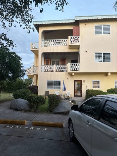 The image depicts the exterior of a three-story residential or commercial building in Olde Yard Village, British Virgin Islands, under an overcast sky. The building is painted light cream or yellow and features multiple balconies with white decorative cross-hatch railings. Several windows are visible, some fitted with white louvered security bars, and others on the second story have open wooden shutters. A light blue-green decorative trim runs along the roofline.
On the ground level, a dark brown door with glass panes serves as the main entrance, accessed via a paved pathway. To the left of this entrance, two white plastic chairs, one dark metal chair, and a closed blue and white striped umbrella are arranged on a small patch of grass. The immediate foreground includes a paved parking area with two wooden wheel stops and the word "VISITORS" painted in white on the asphalt. A light-colored sedan or hatchback is partially visible on the right, parked in the foreground.
The environment surrounding the building includes manicured green grass, various green and reddish-purple shrubs, and several large grey boulders. Dark green tree foliage is visible in the upper left foreground, and distant green hills with additional trees can be seen in the background on the left. No individuals are present or engaged in any actions within the frame.