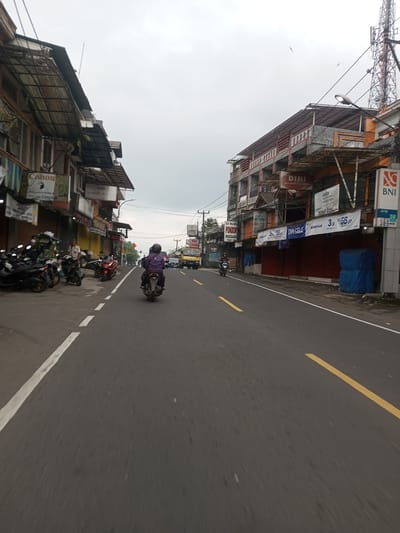 The image captures a street scene in what appears to be an urban or semi-urban area. A person on a motorbike is driving down the road, away from the camera. On either side of the road, there are buildings of varying heights, predominantly commercial, with signs and awnings. There are other parked motorbikes and shops visible along the roadside. The sky is overcast. Buildings have signs like "Canon", "LAUNDRY DAY MONEY CHANGER," "DINI MINI MARKET&KONGLO," and "BNI ATM". A yellow truck is seen in the distance. The road has a solid white line on one side and broken yellow lines. Overhead electrical or communication lines are visible. It is not possible to determine whether the location is in Taunggyi, Myanmar, as there are no identifying features that confirms that exact location.