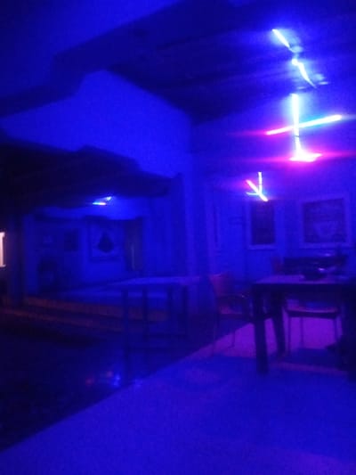 The image depicts a dimly lit interior space, likely a lounge or recreational area, characterized by vibrant blue and purple lighting. In the foreground, a textured blue surface, possibly a carpet or floor, is illuminated by the ambient light, with a diagonal line separating it from a darker, less defined area.
Further into the scene, a dark table with chairs is visible, illuminated by the blue and pink neon lights. The chairs appear to be made of wood and are arranged around the table. To the left of the table, a portion of a wall is visible, with what appears to be framed artwork or pictures hanging on it. One frame shows a dark, abstract shape, while another is less discernible due to the lighting.
Above the table, neon lights are prominently displayed. One set of lights forms a vertical structure with horizontal crossbars, emitting a bright white and pink glow. Another set of lights, also in white and pink, appears to be suspended from the ceiling, with one section forming an angular shape. The ceiling itself is dark and textured, with shadows cast by the overhead lighting.
The overall atmosphere is one of a modern, possibly entertainment-focused establishment, with the neon lighting creating a dynamic and somewhat abstract visual effect. The scene is devoid of any visible individuals. Based on the context provided, this scene is located in Sabon Gari, Nassarawa, Nigeria.