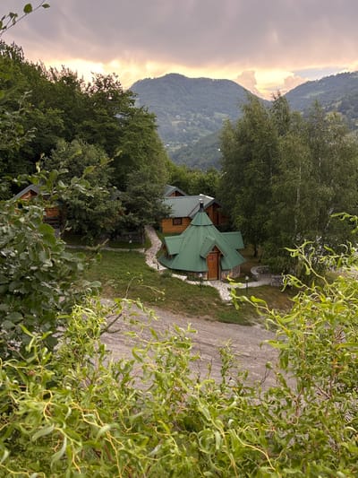 The image depicts a scenic view of a wooden cabin nestled within a lush, mountainous landscape in Mekića Kula, Montenegro. The cabin, with its distinctive green roof, is surrounded by dense trees. A winding path, made of stone and gravel, leads to the entrance of the cabin.
In the foreground, the branches of a leafy tree partially obscure the view, adding a natural framing to the scene. The background is dominated by rolling hills covered in verdant foliage, leading up to a majestic mountain range. The sky is partly cloudy, with the sun breaking through in the distance, casting a soft, warm glow over the mountains. The time of day appears to be late afternoon or early evening, judging by the quality of light.
The overall atmosphere is serene and tranquil, evoking a sense of peace and connection with nature. The location's rural and mountainous setting is emphasized by the natural elements and the rustic architecture of the cabin. There is no visible text in the image.