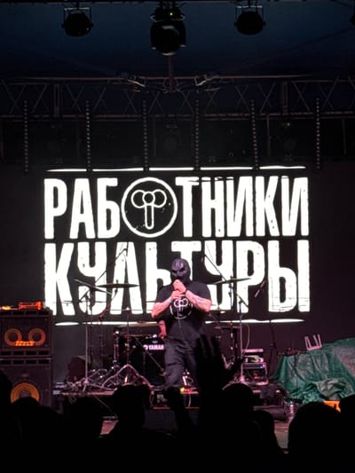 The image depicts an indoor live music performance in Большая Кивара, Russia. Centered on the stage, a performer wearing a dark mask and a dark t-shirt with a distinct circular logo is holding a microphone, actively engaging in the performance. The logo features a central vertical line with two symmetrical loops at its top. Behind the performer, a large illuminated screen displays the Cyrillic text "РАБОТНИКИ КУЛЬТУРЫ" (Workers of Culture) in bold white letters, with the aforementioned circular logo incorporated into the first word.
Stage equipment visible includes a drum kit with a "YAMAHA" branded kick drum behind the performer, and two stacked speaker cabinets with illuminated circular patterns on their grilles positioned to the viewer's left. Various microphone stands, cables, and electronic devices are scattered across the stage. To the viewer's right, a green tarp covers stage equipment, with a white, U-shaped object resting on it. Overhead, metal truss structures support dark stage lights and other rigging. In the immediate foreground, a crowd of audience members is silhouetted, with some hands raised towards the stage. The overall lighting is dim, emphasizing the illuminated screen and the performer.