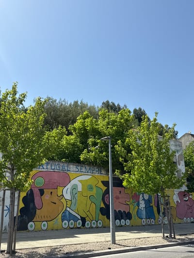 The image shows a mural in Torres Vedras, Portugal, depicting colorful, cartoon-like figures on a wall. The word "PORTUGAL" is vertically oriented to the left of the figures, along with "SEMPRE" underneath. There are trees and foliage partially obscuring the mural. A utility pole stands in front of it. A narrow strip of street is visible to the right. The sky is clear and blue.