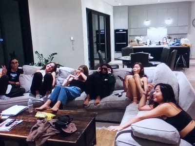 The scene is an interior living space in Aktash, Uzbekistan, featuring an open-concept living room and kitchen area. Six young females are present, casually arranged on a large light-gray L-shaped sectional sofa.
From left to right:
1. A female with dark hair and glasses wears a black top and dark pants, seated with legs crossed on the leftmost section of the sofa, making a "peace" sign with her left hand.
2. A female lies on her side on the sofa's backrest, wearing a white top and dark pants.
3. A female is seated upright on the sofa, wearing a black top and blue jeans, with her bare feet on the sofa. Her hands are positioned near her face.
4. A female is seated upright, wearing a dark hoodie or top and sunglasses, holding a remote control in her right hand. Her bare feet are on the sofa.
5. A female is seated with legs crossed, wearing a light-colored, possibly beige, top, holding a mobile phone or remote in her right hand. Her bare feet are visible.
6. A female is positioned on the rightmost section of the sofa, leaning on her left arm, wearing a black tank top, and looking towards the center of the group.
In front of the sofa, a rectangular wooden coffee table holds a clear plastic water bottle, a yellow snack bag (chips), a crumpled brown fabric item (possibly a sweater or blanket), a black sling bag, a brown cap, a TV remote control, and several magazines or papers. A potted plant is visible behind the sofa on the left.
The background reveals a modern kitchen with light-gray upper cabinets, dark lower cabinets, and a dark island countertop. Two circular pendant lights hang above the island. A black built-in double oven unit is visible on the left side of the kitchen. A round dining table with black chairs is situated to the left of the kitchen island. White flowers in a vase and other miscellaneous items are on the kitchen island. The walls are light-colored, and the flooring appears to be light-toned wood.