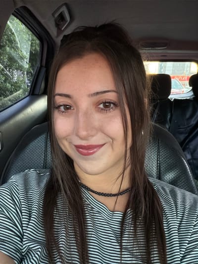 A young woman with brown hair smiles. She's wearing a black and white striped shirt and a beaded necklace. Her eyes are brown and she has light freckles. The photo is taken inside a car, with the car's interior and a glimpse of the outside visible in the background. The image is rotated counterclockwise.