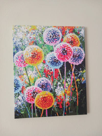 A brightly colored painting of dandelions hangs on a light-colored textured wall. The painting depicts a cluster of dandelions in various stages of bloom, rendered in bold, abstract brushstrokes of pink, orange, yellow, blue, and white. The dandelions are set against a backdrop of green foliage and hints of other small flowers. The painting is presented in a landscape orientation. The location is Mamaivtsi, Ukraine.