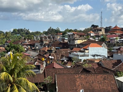 The image captures a dense urban landscape of Klungkung, Indonesia, characterized by a multitude of terracotta-tiled roofs that dominate the foreground and midground. Buildings are tightly packed, with varying architectural styles, including traditional Balinese structures featuring ornate elements and more modern concrete buildings. Lush green tropical foliage, including palm trees with prominent fronds in the lower left, intersperses the built environment, suggesting a warm, humid climate. In the distance, a prominent cellular tower stands against a partly cloudy blue sky. The scene depicts a vibrant, possibly residential and commercial area, with no visible individuals or specific activities taking place. The overall impression is one of a well-established, bustling Indonesian town or city.