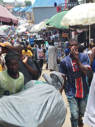 The image captures a crowded marketplace scene in Suleja, Nigeria. People are densely packed, and many vendors have set up stalls with goods covered by umbrellas for shade. One man wears a blue and red soccer jersey, holding his hand near his face. There are various goods on display at the stalls, including boxes of sugar. Many people are looking directly at the camera.