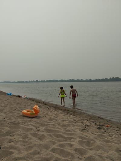 The image depicts a sandy beach along a wide river under an overcast sky. Two children are standing at the edge of the water, with one child, a girl wearing a neon yellow swimsuit, wading in the shallow water. Next to her, a boy in red swim trunks is also standing in the water. In the foreground, an orange inflatable toy shaped like a duck rests on the sand, and further up the beach, other toys and bags are visible. The background shows a treeline across the river. The scene is likely set in the vicinity of Bibikha, Russia, a village known for its river access.
