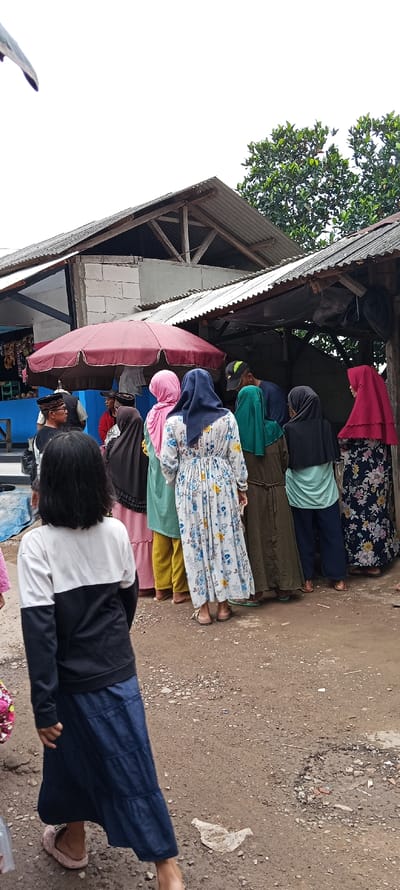 The image shows a group of people standing outside a small, open-sided store. There are 8 people in total, including a young girl with black hair in a white and black sweater and a long blue skirt who is standing near the bottom of the frame. The girl is facing slightly to the right, and her left leg is extended forward, while her right leg is bent at the knee. Her left foot is resting on the ground, while her right foot is off the ground. 

The people are standing in a line, facing the store. The woman in the front of the line is wearing a white floral dress and has a pink scarf on her head. The woman behind her is wearing a green dress.  The other people in the line are wearing various clothing, including headscarves and traditional clothing. 

The store has a corrugated metal roof and wooden support beams. There is a red umbrella on the left side of the store. The image is likely taken in Kecamatan Kutawaringin, Indonesia, based on the coordinates provided. The ground is rough and dusty, and there are some pieces of trash on the ground. The scene is typical of a bustling, everyday market scene in Indonesia.