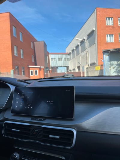 The image presents an urban exterior scene observed from the interior of a vehicle, located in the city of Sel’khoztekhnika, Russia. The immediate foreground shows a dark gray vehicle dashboard featuring a wide, rectangular digital infotainment display. The active screen exhibits a dark interface with white text, indicating "Bluetooth" connectivity, current time "10:41," and temperature "20.0°C." Russian text relating to audio playback, such as "Не плачь" and "Бульдог," is also visible. Below the screen, horizontal air vents with chrome accents and a red hazard light button are present. A brushed metallic trim panel extends across the passenger side of the dashboard. A small black mounting device is affixed to the inner windshield.

Through the windshield, a clear blue sky is visible above. The background comprises multiple structures. To the left, a multi-story building constructed of orange-red brick features several white-framed windows, with a smaller, single-story red-brick structure at its base. To the right, a building with a gray, corrugated metal facade and covered windows stands adjacent to a section of red-brick wall containing white-framed windows. Below this brick section is a dark blue corrugated metal gate or door. An elevated, enclosed skywalk or bridge, constructed of light gray, possibly corrugated metal with multiple windows, connects structures in the middle distance. A dark brown building section is partially visible between the left brick building and the skywalk. Overhead utility wires traverse the sky. A light gray street lamp extends from the right-side building. No individuals are present within the scene.