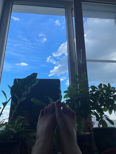 The image shows a close-up view of a person's feet resting on a windowsill, with several houseplants in the foreground. Through the window, a bright blue sky with white clouds is visible, suggesting it is daytime. The overall scene appears to be a domestic setting, possibly an apartment or house, located in the city of Cherepovets, Russia.