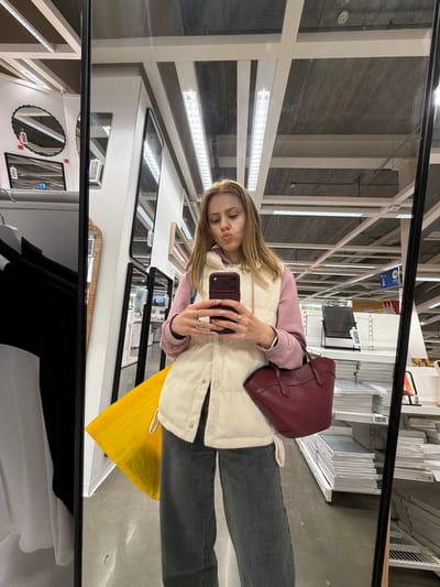 A young woman is taking a selfie in a store, reflected in a large mirror. She is wearing a pink hoodie and a cream-colored corduroy vest, dark gray wide-leg pants, and rings on her fingers. She holds a maroon handbag in her right hand and a smartphone in her left. A bright yellow bag is visible behind her. The background shows shelving units with various items, fluorescent lighting, and architectural elements of a retail space. The overall scene suggests a shopping environment, possibly in an IKEA store given the typical retail display. The location is identified as Son en Breugel, Netherlands.