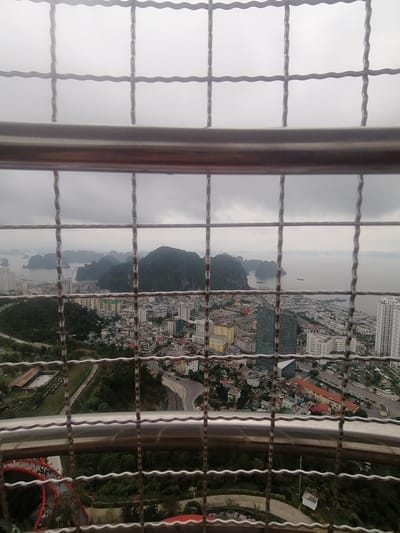 View of Hong Gai Ward, Vietnam, through a safety barrier. Buildings and a bay with islands visible. Overcast sky. A red bridge is seen in the foreground below.