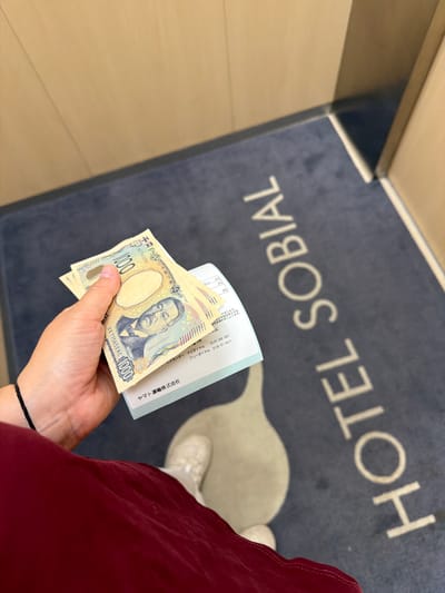 A first-person perspective photograph captures a person's hand holding two Japanese 1000 Yen banknotes and a white receipt, standing on a dark blue doormat with white lettering. The hand is light-skinned, with a thin black bracelet on the wrist. The lower part of a red garment is visible on the left, and a light-colored shoe is partially visible on the bottom right. The 1000 Yen banknotes feature the portrait of Hideyo Noguchi; a partial serial number "AA177652NC" is visible on one note. The white receipt, held beneath the currency, displays Japanese text, including "ヤマト運輸株式会社" (Yamato Transport Co., Ltd.), amounts in Yen, and customer service contact information: "サービスセンター ナビダイヤル 0570-200-000" and "フリーダイヤル 0120-01-9625". The doormat clearly spells out "HOTEL SOBIAL" in capital sans-serif letters, with a stylized white teardrop-shaped emblem positioned beneath the 'L' of 'HOTEL'. The surrounding environment consists of light brown or beige walls, possibly wood-paneled or painted. This scene is located within a Hotel Sobial, definitively in Osaka, Japan, given the Japanese currency, language on the receipt, and the presence of Yamato Transport.