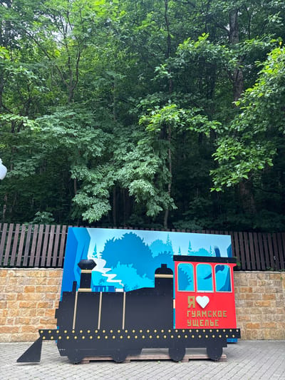 This image features a colorful, cartoonish cutout of a train, designed as a photo opportunity. The train has a red engine with three arched windows, through which silhouettes of birds are visible. A white heart is placed next to the text "Я ГУАМСКОЕ УЩЕЛЬЕ", which translates to "I love Guamka Gorge". The rest of the train is black with yellow accents, resembling the wheels and rivets.

The train cutout stands in an outdoor setting. To the left of the train, lush green trees with dense foliage create a natural backdrop. To the right, a rustic wooden fence and a stone wall are visible, hinting at a park or an attraction area. The ground is paved with gray interlocking stones.

The scene appears to be set during the daytime, with natural light illuminating the area. The weather seems pleasant, as indicated by the vibrant greenery. The overall impression is one of a tourist attraction aimed at creating a fun and memorable experience for visitors in Guamka, Russia.