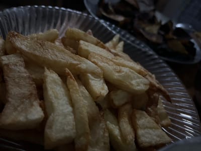 A close-up captures a clear bowl overflowing with thick-cut, golden-brown french fries. Some fries exhibit darker, crispier edges. The bowl's ridged design adds a subtle texture to the presentation. Another bowl, partially visible in the lower left corner, contains darker-colored fries. The image is somewhat dark.