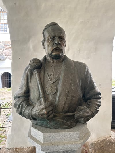A bronze bust of a man with a mustache stands on a granite pedestal. The man is wearing a jacket with a medal around his neck and is holding a mace in his right hand. The bust is located in Solovetskiy, Russia, specifically on the grounds of the Solovetsky Monastery. The background shows a white stucco wall with arched windows and stone buildings.