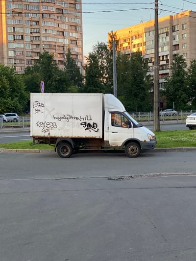 A white panel van with graffiti on its side is parked on the right side of a street in Ladozhskoe Ozero, Russia. The van is facing right and is partially obscured by a grassy median. The graffiti appears to be in black and white spray paint and includes various words and symbols. In the background, there are apartment buildings, trees, and a power pole. The sky is clear and the lighting suggests it is either early morning or late afternoon. The street is paved with asphalt and shows some signs of wear and tear, including cracks and potholes.