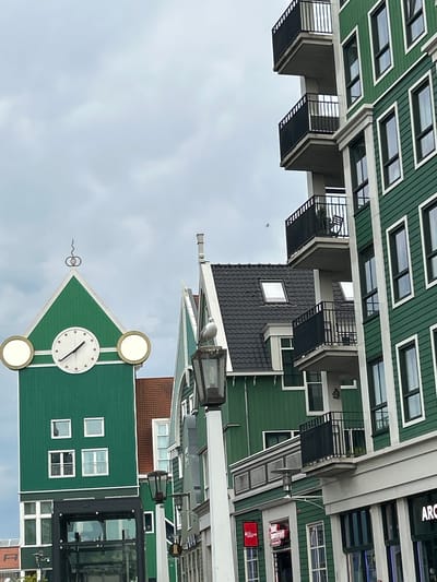 The image depicts a street scene in Zaandam, Netherlands, featuring distinctive green buildings with white trim. In the foreground, a tall, green tower with a white-faced clock is prominently visible. The clock has black hands and two circular, illuminated elements on either side. Below the clock, the tower has several square windows, and at its base, a glass-enclosed structure with metal framing suggests an entrance or elevator. To the right of the tower, a modern, multi-story building with a green facade and multiple balconies with black railings rises. Its windows are rectangular with white frames. The background reveals more of the green buildings, some with dark, pitched roofs and dormer windows, as well as a glimpse of a red-tiled roof. A street lamp with a seagull perched on top stands between the green tower and the modern building. The sky is overcast with diffused light. The scene appears to be from a public area or a commercial district, with shopfronts visible on the ground floor of the modern building, including signage that reads "ARC". The architecture is characteristic of the Zaanse Schans area, known for its preserved historical buildings and modern interpretations thereof.