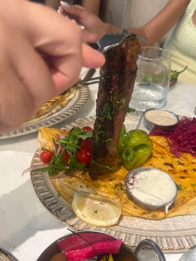 A close-up, high-angle shot shows a hand holding a fork that is spearing a grilled meat kebab. The kebab is brown and appears to be seasoned with herbs. It is served on a flatbread with a variety of accompaniments including a green bell pepper, cherry tomatoes, red onion, and leafy greens. There are also two small metal cups containing white dips, one sprinkled with red spice. A slice of lemon is visible on the side. The meal is presented on a decorative plate, which sits on a white, marble-like surface. In the background, a glass of water and a green chili pepper are visible, as are parts of other dishes and people's hands and arms. The scene suggests a restaurant setting, likely in Aventura, Florida, USA.