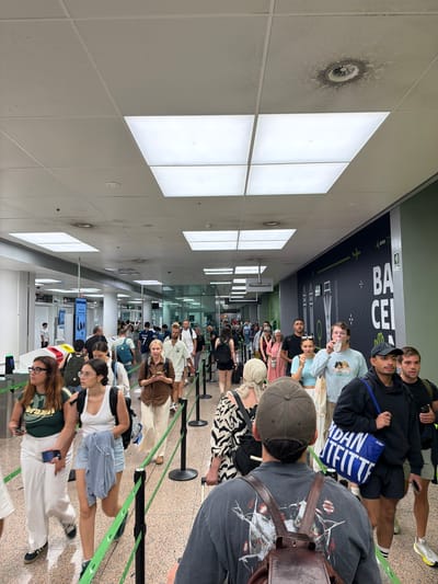 The image depicts a busy indoor scene, likely an airport terminal, filled with people in a queue. The environment features a long, bright hallway with a drop ceiling containing fluorescent light panels. Along the right wall is a large dark green banner with white text and graphics, including the word "BARCELONA". People of various ages and ethnicities are visible, many carrying luggage and wearing casual attire. They are moving in the same direction, suggesting they are proceeding through a checkpoint or to a gate. The foreground shows several individuals in close proximity, with some looking forward and others engaged with their phones. Green stanchion ropes are set up to guide the flow of people. The ceiling shows signs of wear, with a circular discoloration and some debris around a vent. The overall impression is one of crowded transit within an airport.