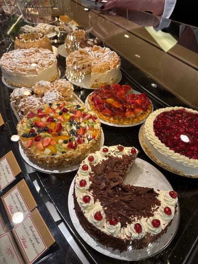 The image captures a display case filled with various cakes and pastries, likely within a bakery or café in Cochem, Germany. The cakes are arranged on plates and include Black Forest cake (Schwarzwälderkirsch), strawberry cake (Erdbeertorte), raspberry yogurt cake (Himbeerjoghurt), and other fruit-topped cakes. Price tags are visible below the cakes, indicating that the cakes are for sale. A portion of a person's arm, possibly a store clerk, is visible on the left side of the image. The reflective glass of the display case is visible.