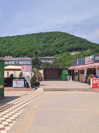 The image shows a street scene in Dzhubga, Russia, with a verdant, tree-covered mountain in the background. In the foreground, there are several kiosks or small shops. One kiosk on the left is painted green and yellow, with an ice cream display featuring colorful cones and tubs. Next to it, a more prominent kiosk has a sign that reads "УЮТНОЕ МЕСТО" (Cozy Place) with a location pin icon. This kiosk displays pictures of food, including what appear to be burgers and fries. Adjacent to this, another establishment has a striped awning and a sign that says "СТОЛОВАЯ" (Canteen). Further to the right, a red sign with white lettering reads "АПЕ" which is likely the beginning of a word like "АПЕТИТ" (Appetite) or "АПТЕКА" (Pharmacy), followed by a Russian letter that could be "К". The ground is paved with a patterned tile design in some areas and regular paving stones in others. There are also some potted plants and trees, including a flowering bush with pink flowers on the right side. The sky is partly cloudy and bright. No people are clearly visible in the scene.