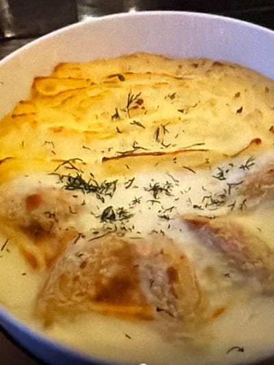 The image shows a close-up of a white bowl containing a baked dish. The dish appears to have a golden-brown crust on top, which is textured and slightly uneven. Scattered across the crust are small green specks, possibly herbs like dill. Beneath the crust, some creamy sauce and pieces of food are visible. The food appears to be light brown in color and somewhat rounded. The focus is on the texture and appearance of the food, with the background being blurred and dark, suggesting indoor lighting. There is no indication of the city of Lopatino, Russia, or any people in the image.