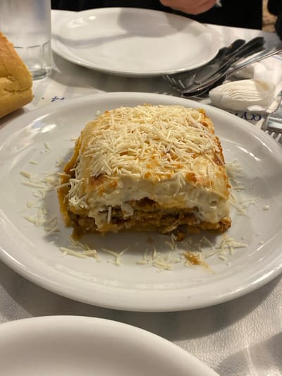 A rectangular portion of moussaka, featuring layers of minced meat, vegetables, and a thick béchamel sauce, is centered on a round white ceramic plate. The dish is heavily garnished with shredded white cheese, with some strands scattered on the plate around its base. The plate rests on a white textured tablecloth. In the upper left background, a clear glass of water and a light brown bread roll are visible. To the upper right, an empty white plate, a stack of silver cutlery including forks and knives, and a folded white napkin with a partial blue inscription are arranged. The top edge of the image shows dark fabric, indicating the presence of a person. This scene takes place in a dining establishment in Megalochori, Greece.