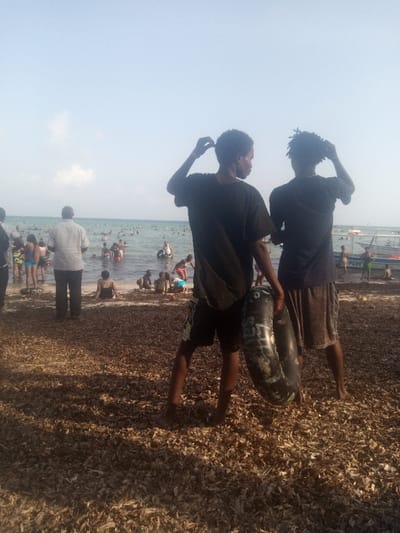 This image depicts a crowded coastal scene in Shanzu Ward, Kenya, under a clear, light blue sky with minimal cloud cover.
The foreground consists of a beach covered extensively with a layer of dried, brown organic matter, likely seaweed or plant debris. Visible shadows suggest sunlight originating from the right side of the frame. Two young males stand in the immediate foreground, observed from behind, facing the ocean. The male on the left wears a dark t-shirt and dark shorts, with his right hand raised to his head, appearing to adjust his hair. The male on the right wears a dark t-shirt and brown shorts, also with his left hand raised to his head, adjusting his hair. He holds a dark, possibly deflated or partially deflated inflatable ring with visible white markings against his left leg.
In the midground, a diverse group of individuals, including adults and children, occupies the shoreline and the shallow, light blue-green water. One adult male, seen from behind, stands prominently in a light plaid shirt and dark trousers. Several people are wading, sitting, or playing in the water near the shore. Many individuals in the water are utilizing inflatable rings.
Further into the background, a large number of people are visible submerged in the water. Several small watercraft, likely boats, are present near the horizon line on the right side of the image. The overall environment suggests a recreational beach setting with numerous people engaged in water activities.