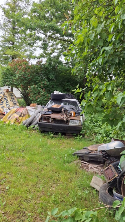 An abandoned, rusty car is parked on overgrown grass in a rural area, possibly Timmolovo, Russia, surrounded by various discarded items. The car's hood is open, revealing an engine with exposed parts, and the windshield is cracked. Several tires are stacked on the car's roof, and other automotive parts, along with assorted debris, are scattered around the vehicle. In the background, dense foliage and trees create a natural backdrop, with a partially visible building suggesting a residential area nearby. The scene is static, with no visible activity or people.