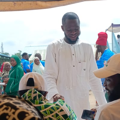 The image depicts a man wearing a white, long-sleeved traditional garment and smiling in what appears to be an outdoor gathering. The man is in the foreground, surrounded by a crowd of people with a mix of casual and traditional clothing. Some people are wearing green and white outfits, others blue shirts with political party logos. One person wears a red hat. There are buildings in the background. A partial structure provides shade from above. The environment suggests a daytime political or community event. There is no specific information in the image to confirm that the location is Gudi, Nigeria.