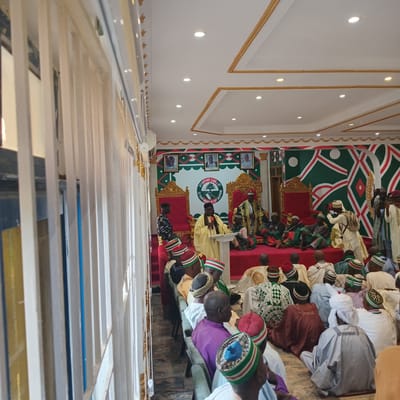 The image depicts an indoor gathering, likely a ceremony or formal event, taking place in Unguwar Gaya, Nigeria. A man in a light yellow robe stands at a podium, speaking into a microphone. He is positioned on a raised platform draped in red carpet, with two ornate, gold-trimmed thrones behind him. The wall behind the platform features a repeating geometric pattern of green and white, with three framed portraits of men above.
A seated audience fills the space, many wearing traditional Nigerian attire, including caps with red, white, and green bands, and flowing robes. Some individuals are seated on the floor, while others are on rows of chairs. The overall ambiance suggests a community event or a significant cultural occasion. The room has a high ceiling with recessed lighting and decorative gold trim. The foreground is partially obscured by vertical blinds, suggesting the event might be viewed from an adjacent area.