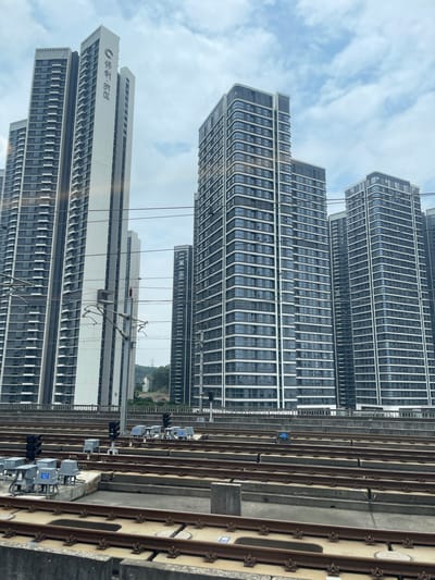 This is a medium shot, taken from a moving vehicle looking out at a modern city scene with residential buildings and train tracks. The buildings are tall, multi-story structures with a repetitive facade of windows and white horizontal accents. The train tracks run parallel to the buildings, with signal lights and electrical equipment visible along the tracks. The sky is overcast with clouds. The image appears to be taken during the daytime.
Visible text: On one of the white buildings, there is Chinese text and a logo. The text appears to be "华润·润园". There are also some small blue and white labels on the signal equipment along the train tracks.
The scene conveys a sense of urban development and infrastructure in Dongguan, China. The image is oriented vertically as if captured through a window.