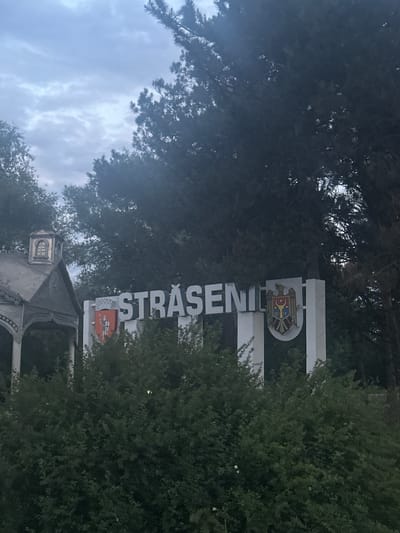 An exterior shot captures a welcome sign for the city of Strășeni, Moldova, under an overcast sky. The sign is large and white with dark lettering spelling out "STRĂȘENI." The city's coat of arms is displayed on the left side of the sign, featuring a red shield with a knight. On the right side of the sign, the national coat of arms of Moldova is visible, a double-headed eagle holding a shield with aurochs head. To the left of the sign is an elaborately decorated, traditional wooden structure with a peaked roof and a small arched window. Dense green foliage, likely bushes and trees, partially obscures the lower portion of the sign and the wooden structure. The overall scene suggests a city entrance or roadside marker.