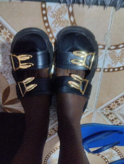 The image provides a downward-facing perspective of a person's lower legs and feet, showcasing a pair of black sandals on a patterned surface.
The central focus is on the footwear. The person is wearing thick-soled, black sandals, possibly with a platform design. Each sandal features two wide, horizontal straps across the top of the foot. A distinctive detail of these sandals is the gold-colored hardware on each strap. These ornaments are shaped like stylized rabbit ears, with two upward-curving, pointed lobes, creating a unique and decorative buckle effect against the dark material of the sandals.
The wearer's legs are visible from just below the knees to the feet. They appear to be covered in dark hosiery or stockings, which gives the skin a smooth, dark, and uniform texture. The natural skin tone, visible through the hosiery, is dark.
The background is a textured, patterned surface, likely a rug, mat, or tiled floor. The base color is a light beige or cream, adorned with intricate designs in a darker brown or golden hue. These patterns include floral or scroll-like motifs, rows of circular dots, and geometric elements such as small square or diamond shapes arranged in lines.
In the lower right corner of the frame, a portion of a blue object is visible, which appears to be a pair of blue flip-flops or open-toed sandals, resting on the same patterned surface.
The lighting seems consistent with an indoor setting, and there are no specific indicators of the time of day. The image appears to be a casual photograph, likely taken to highlight the design of the sandals. There is no discernible text present in the image.