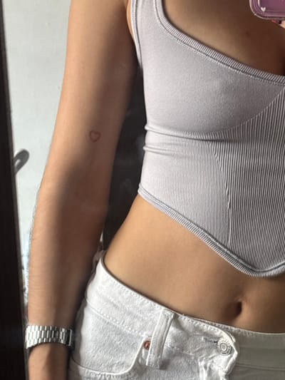 A woman is shown wearing a light gray striped tank top and white shorts. Her arm, adorned with a delicate heart tattoo, rests horizontally. A silver watch is visible on her wrist. The overall composition is a close-up, capturing the body from the chest to the waist, with the focus on the arm and torso.